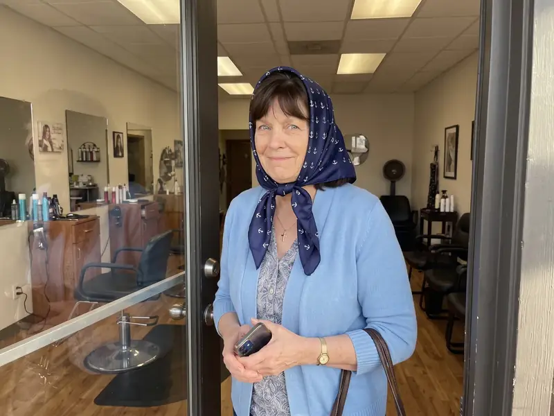 An older woman in a silk scarf standing in a small salon doorway in the morning